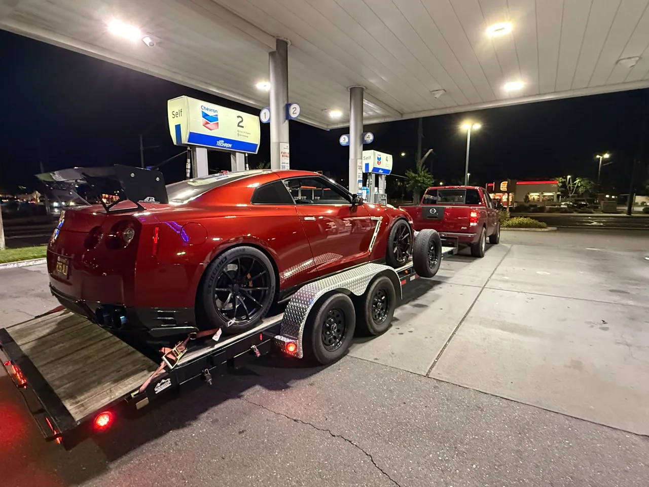 Red performance coupe secured on a trailer at a lit fuel station